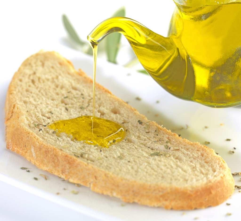 En la imagen se ve una rebanada de pan tostado con una textura crujiente, sobre la cual se está vertiendo un chorro de aceite de oliva virgen extra desde una jarra. El aceite de oliva, de color dorado y brillante, se acumula en el centro del pan, destacando su frescura y calidad. Ideal para representar el uso del aceite de oliva virgen extra en recetas simples y saludables.