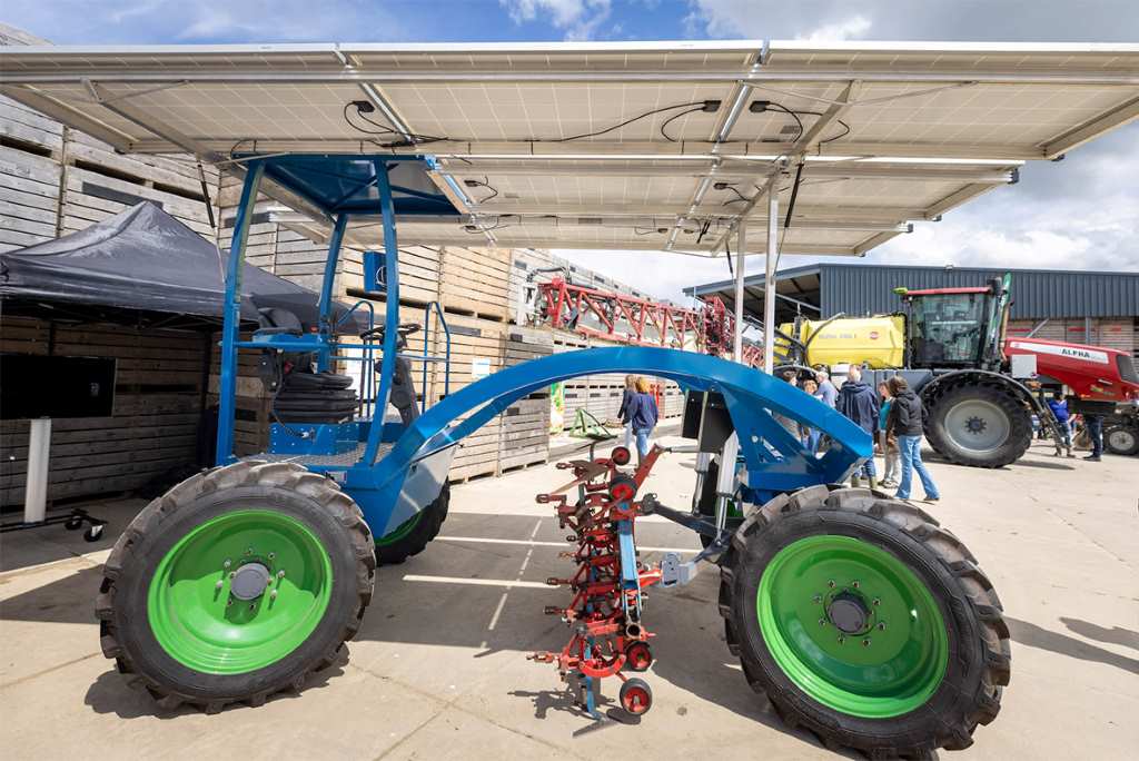 Un tractor solar azul con grandes neumáticos y paneles solares en la parte superior se exhibe en una feria agrícola. El tractor tiene implementos de cultivo en la parte inferior y está diseñado para funcionar con energía solar, ofreciendo una alternativa ecológica a los tractores convencionales. Al fondo, se observan otras máquinas agrícolas y varias personas explorando el área.