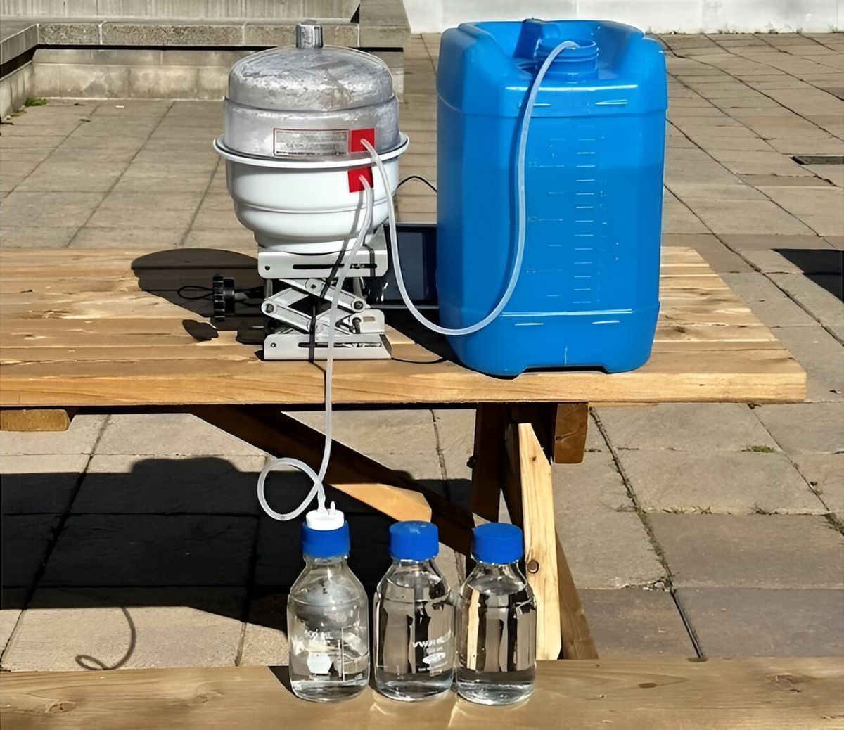 En la imagen, se observa un dispositivo colocado sobre una mesa de madera al aire libre. El sistema parece estar diseñado para desalación o filtración de agua. A la izquierda, hay un tanque cilíndrico blanco con una estructura metálica alrededor, posiblemente un componente clave del proceso. De este tanque sale una manguera que conecta con un contenedor azul más grande, que parece almacenar el agua tratada o a tratar. En la parte delantera, sobre la mesa, se encuentran tres frascos de vidrio transparente con tapas azules, que contienen lo que parece ser agua limpia o desalada, lo que sugiere que el dispositivo ha generado agua potable a partir de agua salada u otro tipo de fuente no potable.

Este conjunto podría ser un prototipo de un sistema de desalación portátil, posiblemente similar al descrito en el artículo, donde se utiliza energía solar para transformar agua de mar en agua potable.