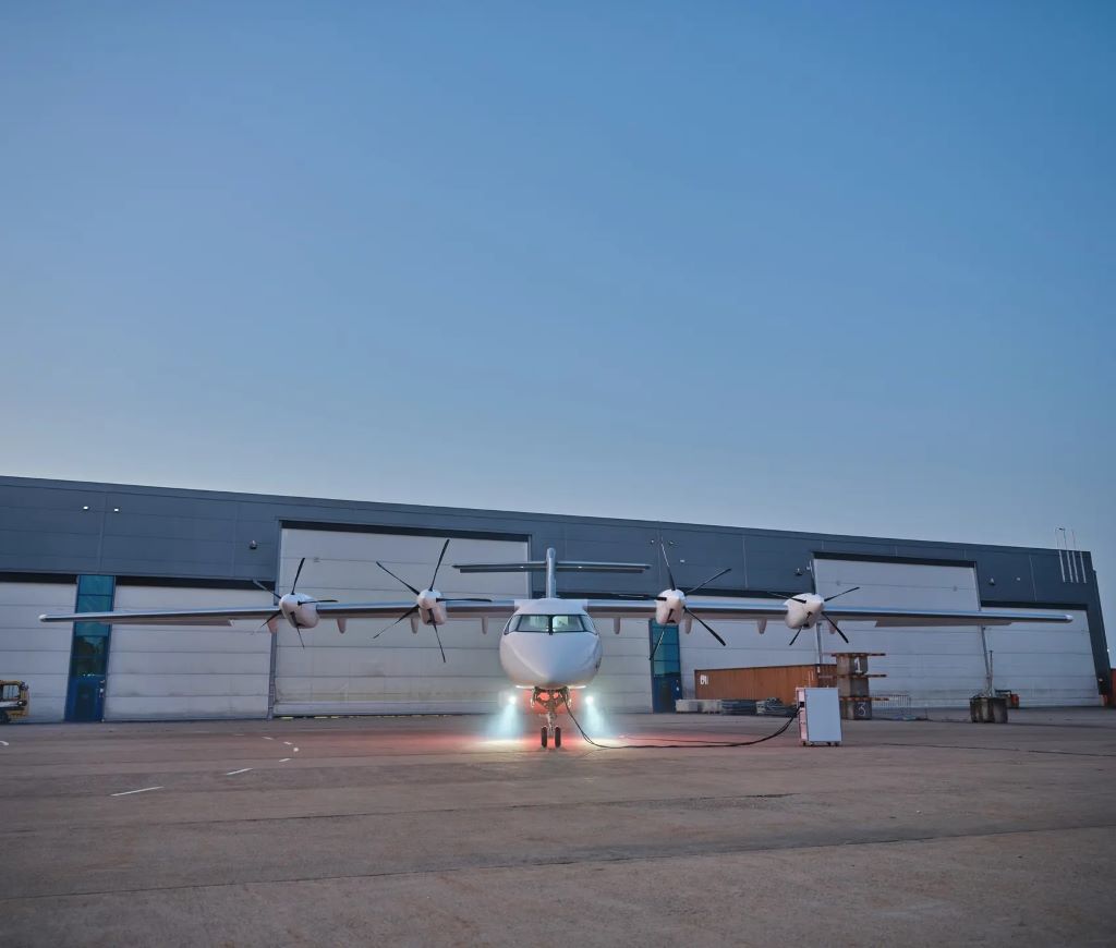 En la imagen se puede observar un avión en tierra, frente a un hangar de mantenimiento, en lo que parece ser un entorno industrial o de aeropuerto. El avión es de color blanco y tiene cuatro hélices: dos en cada ala, situadas a diferentes distancias del fuselaje. Las hélices más cercanas al fuselaje parecen corresponder a los motores eléctricos, mientras que las más alejadas podrían ser las turbopropulsoras adicionales para vuelos de mayor alcance. El entorno muestra algunas señales de vapor o humo cerca del suelo, lo que podría indicar que el avión está en fase de pruebas o preparándose para alguna operación.